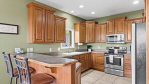 A kitchen with wooden cabinets, a stainless steel refrigerator, a double oven, and a bar area with two stools. - The Greenview Manor - Lake View - 9 Miles from Silver Dollar City (Kimberling City)