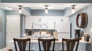 A kitchen with white cabinets, a central island with bar stools, a microwave, stove, and a coffee maker. - Eagle's Nest at The Club- Chic 2BR Golf View Condo (Branson)