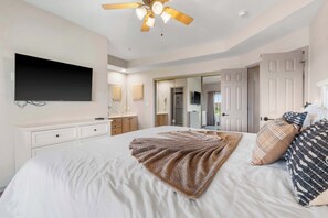 A bedroom with a bed, a ceiling fan, a flat-screen TV, a dresser, a mirror, and a door leading to another room.