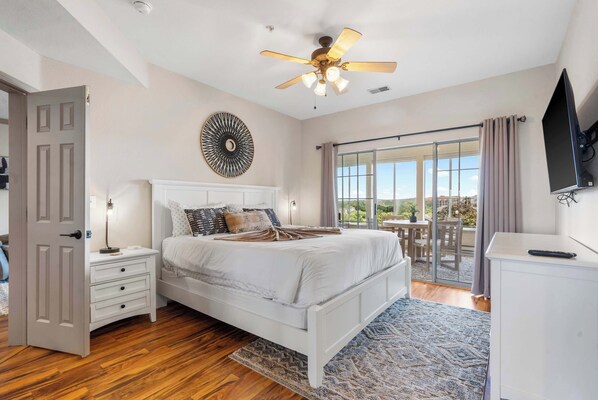 A bedroom with a bed, bedside tables, a ceiling fan, a TV, and a sliding glass door to a balcony.