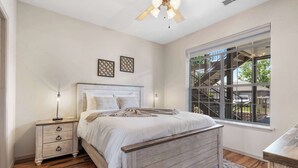Un dormitorio con cama de madera, mesita de noche, lámpara, ventilador de techo y ventana con vista a una escalera y follaje exterior.