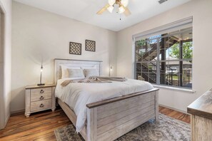 A bedroom with a wooden bed, a nightstand, a lamp, a ceiling fan, and a window with a view of a staircase and greenery outside.