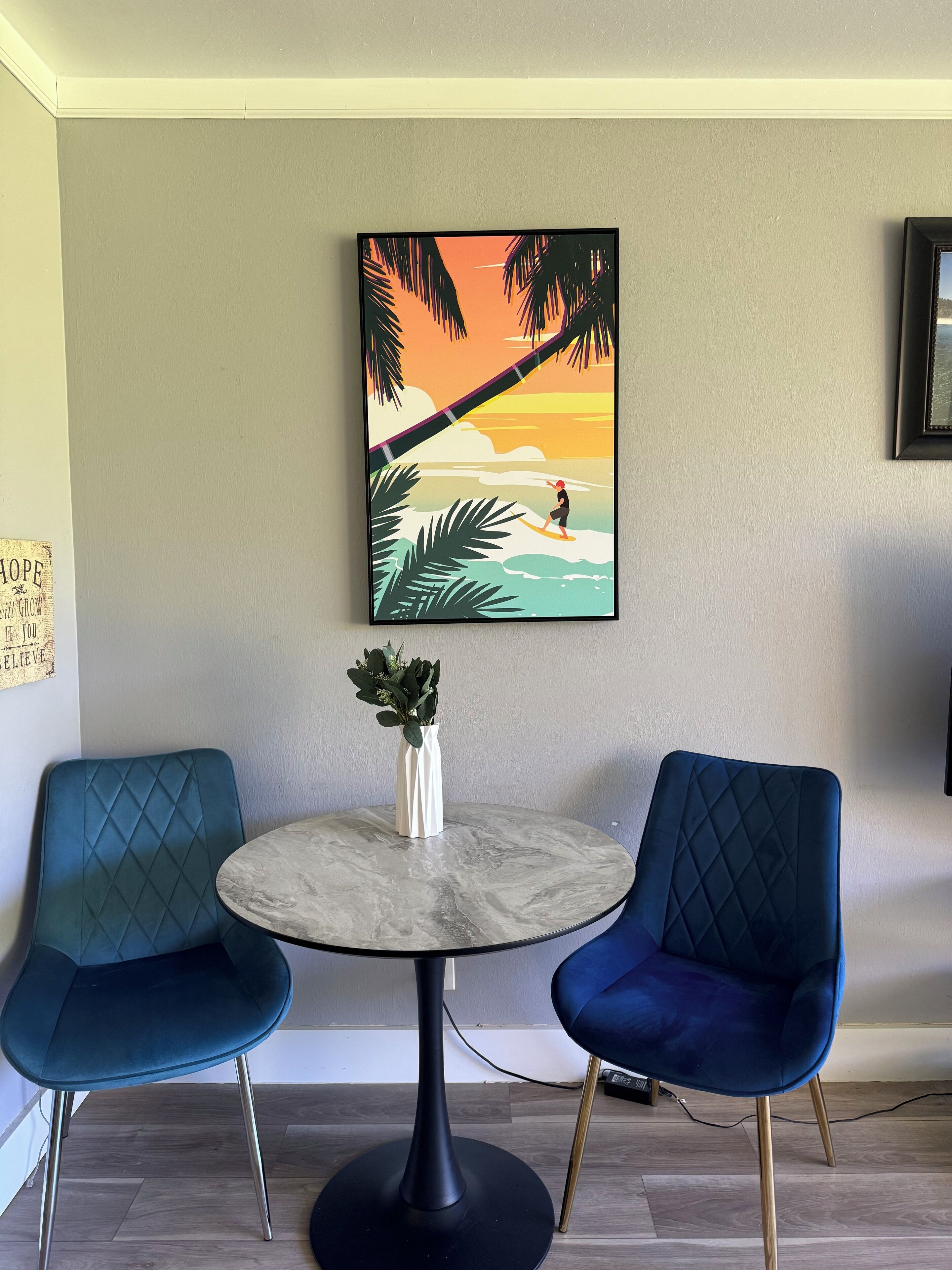 Une pièce avec une table ronde, deux chaises bleues, un tableau représentant un surfeur accroché au mur, et un petit vase blanc contenant des plantes vertes.