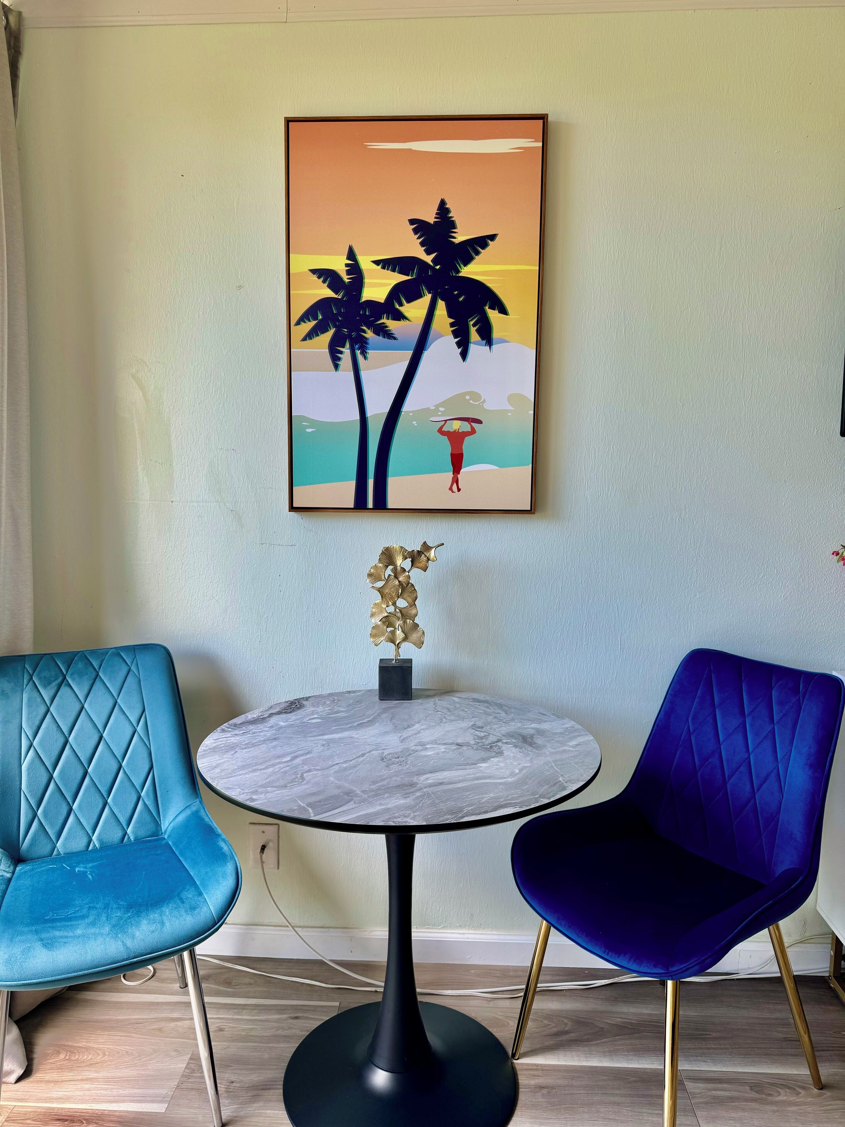 A room with a round marble table, two upholstered chairs, a framed artwork of a beach scene, and a decorative plant.
