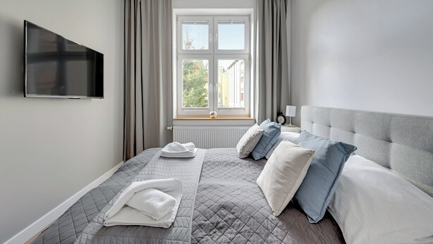 Een moderne slaapkamer met een groot bed, een flatscreen televisie, een raam met gordijnen en een nachtkastje met een lamp.