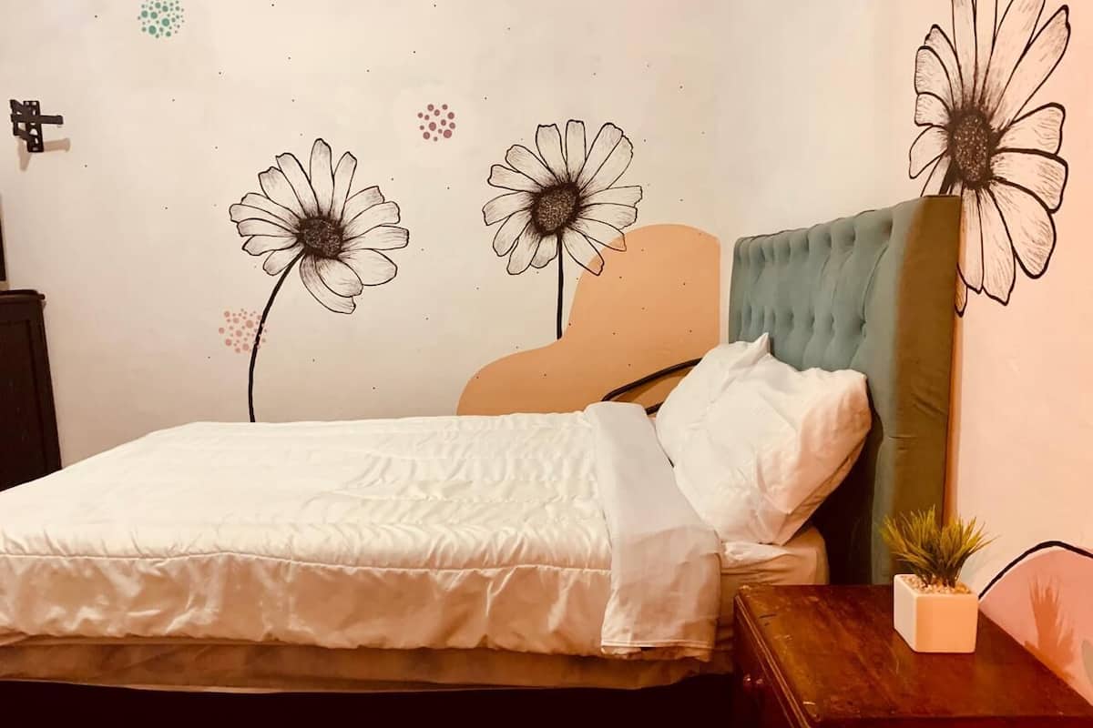 Una cama con edredón blanco, cabecera tapizada en verde y mesita de noche de madera con una planta pequeña.