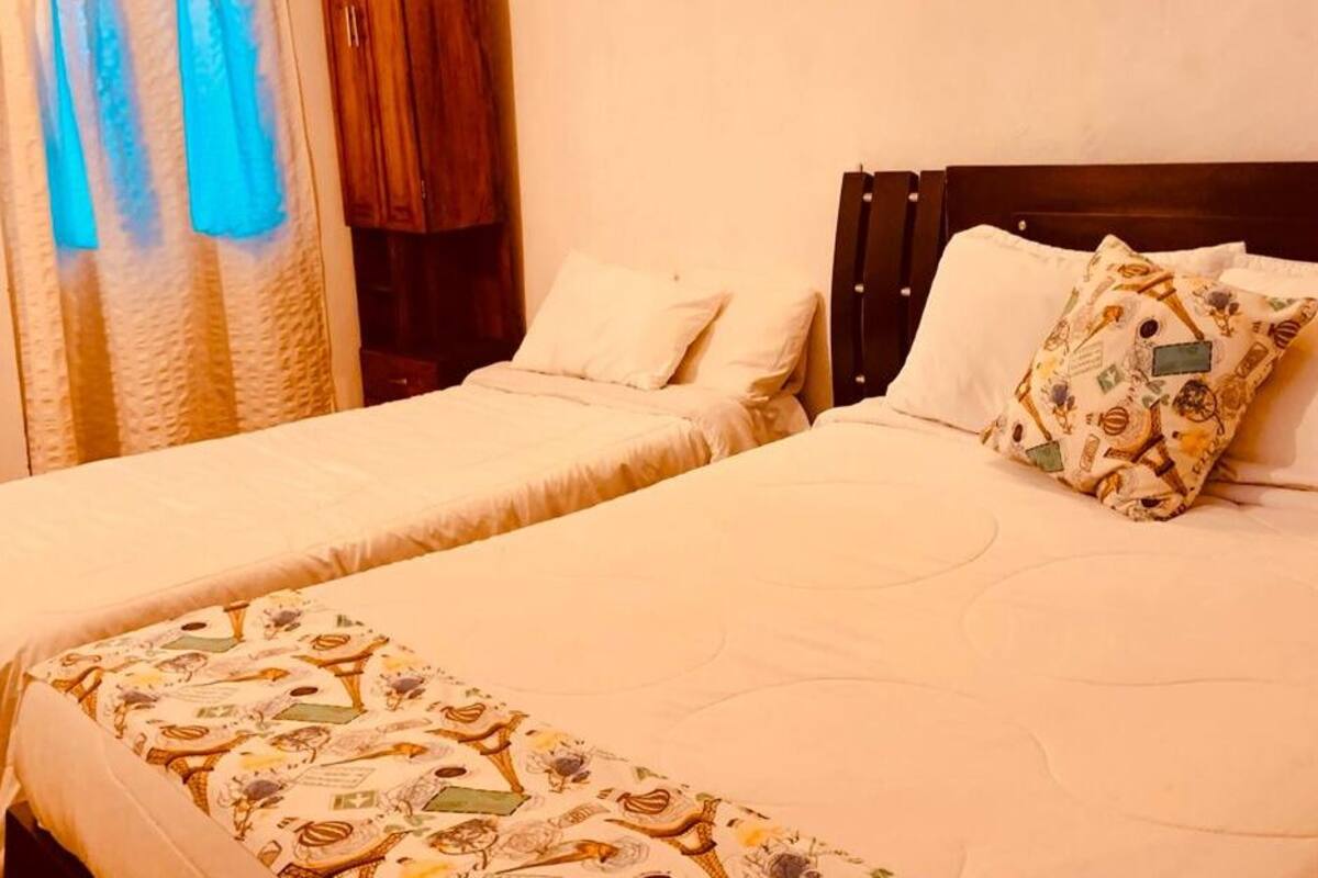 Habitación de hotel con dos camas individuales, cada una con cubrecama y almohadas a juego. La habitación tiene cabecera de madera y cortina con un tono azulado.