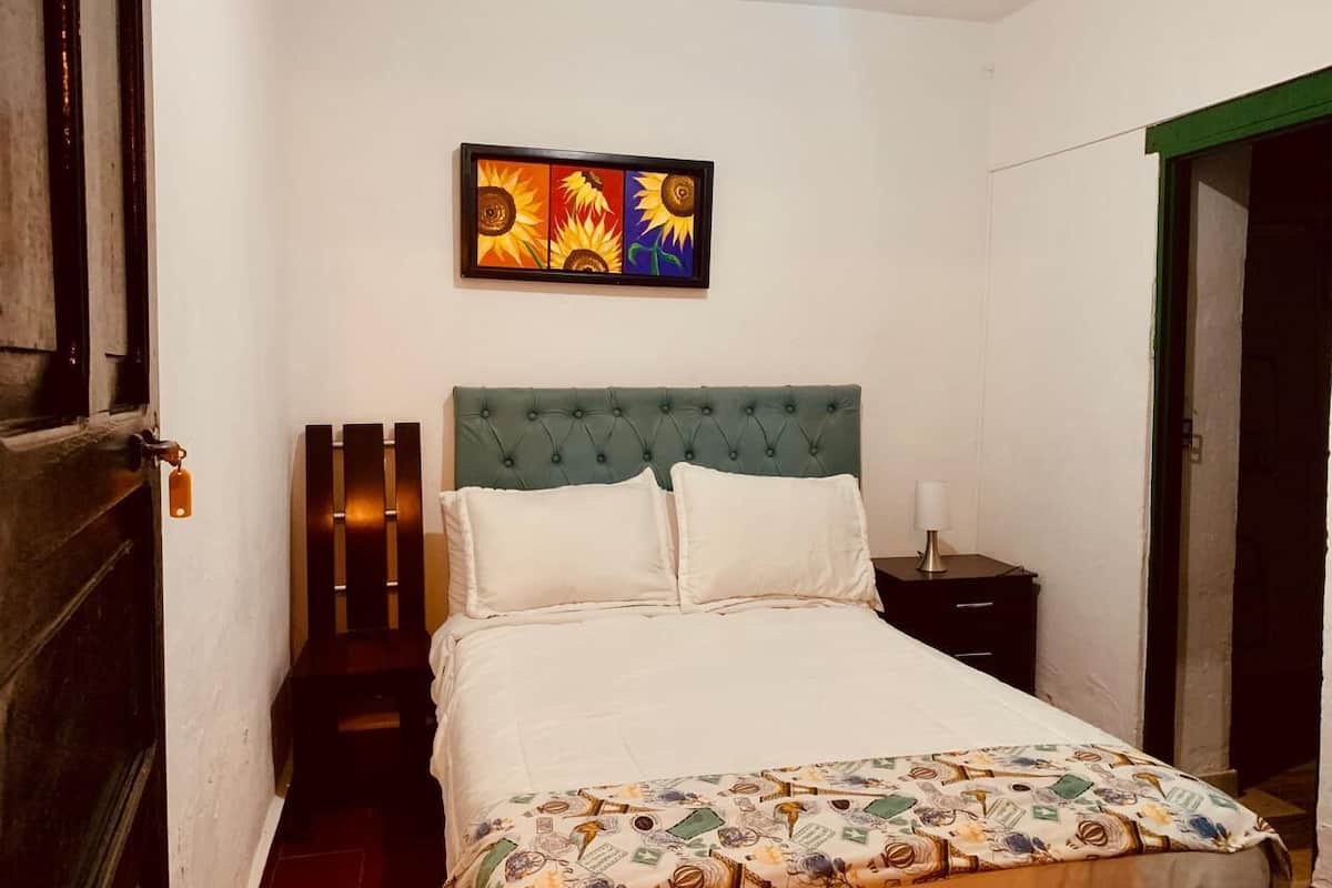 Un dormitorio con cama, mesita de noche, un cuadro en la pared y una puerta.