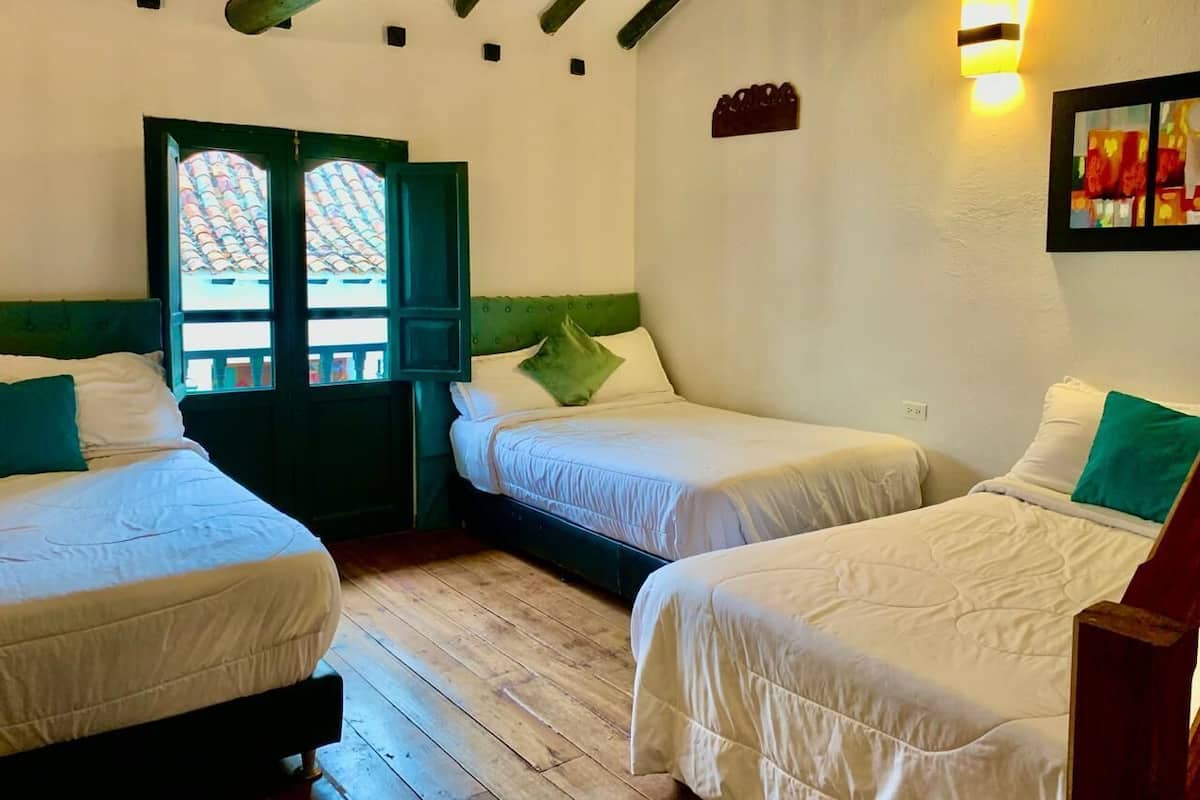 Una habitación con dos camas, piso de madera, una puerta verde y una lámpara fijada a la pared.