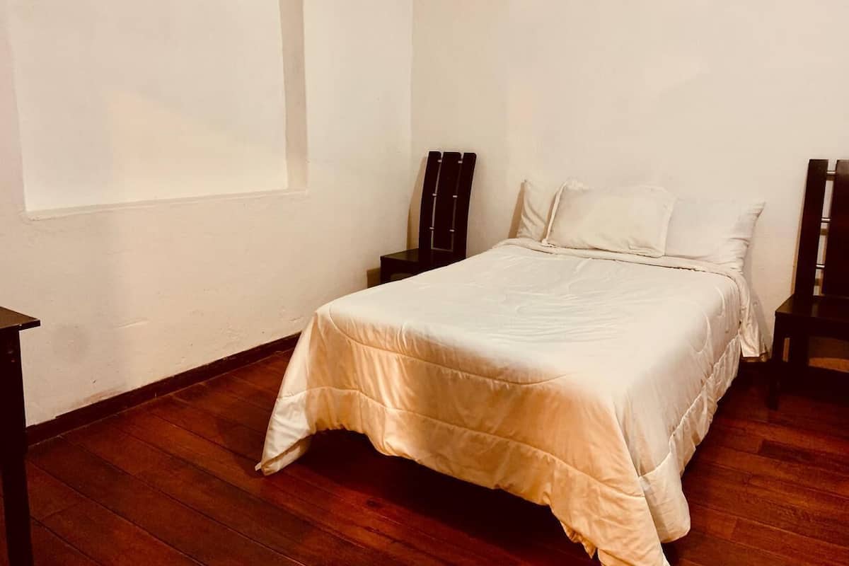 Un dormitorio con cama, suelo de madera, paredes blancas y una ventana.