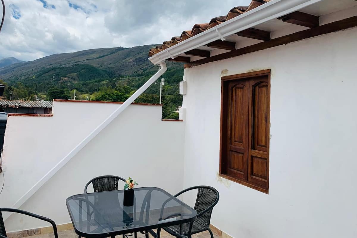Un patio con una mesa y sillas de vidrio, una puerta de madera y vistas a colinas verdes al fondo.