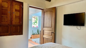 A bedroom with a bed, a wooden wardrobe, a television, and an open door leading to a balcony with plants. - Hotel santone la ville (Villa de Leyva)