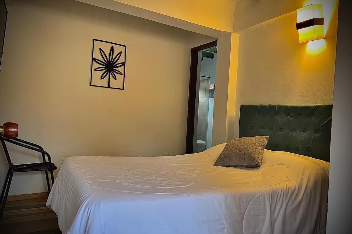 Un dormitorio con cama, una silla, una obra de arte en la pared y una mesita de noche.