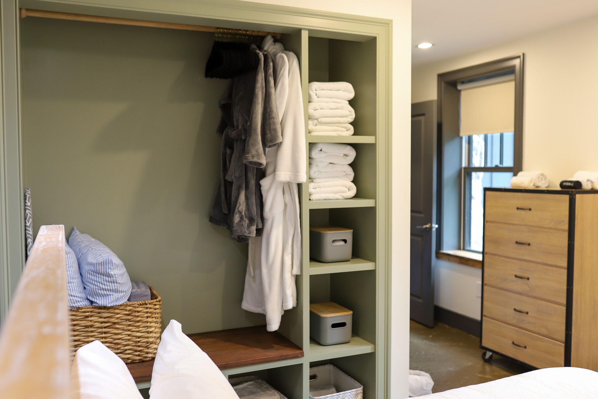 Ein Hotelzimmer mit einem grünen Kleiderschrank, gefalteten Handtüchern, einem Korbwäscher mit Kissen und einer Holzkommode mit Spiegel.