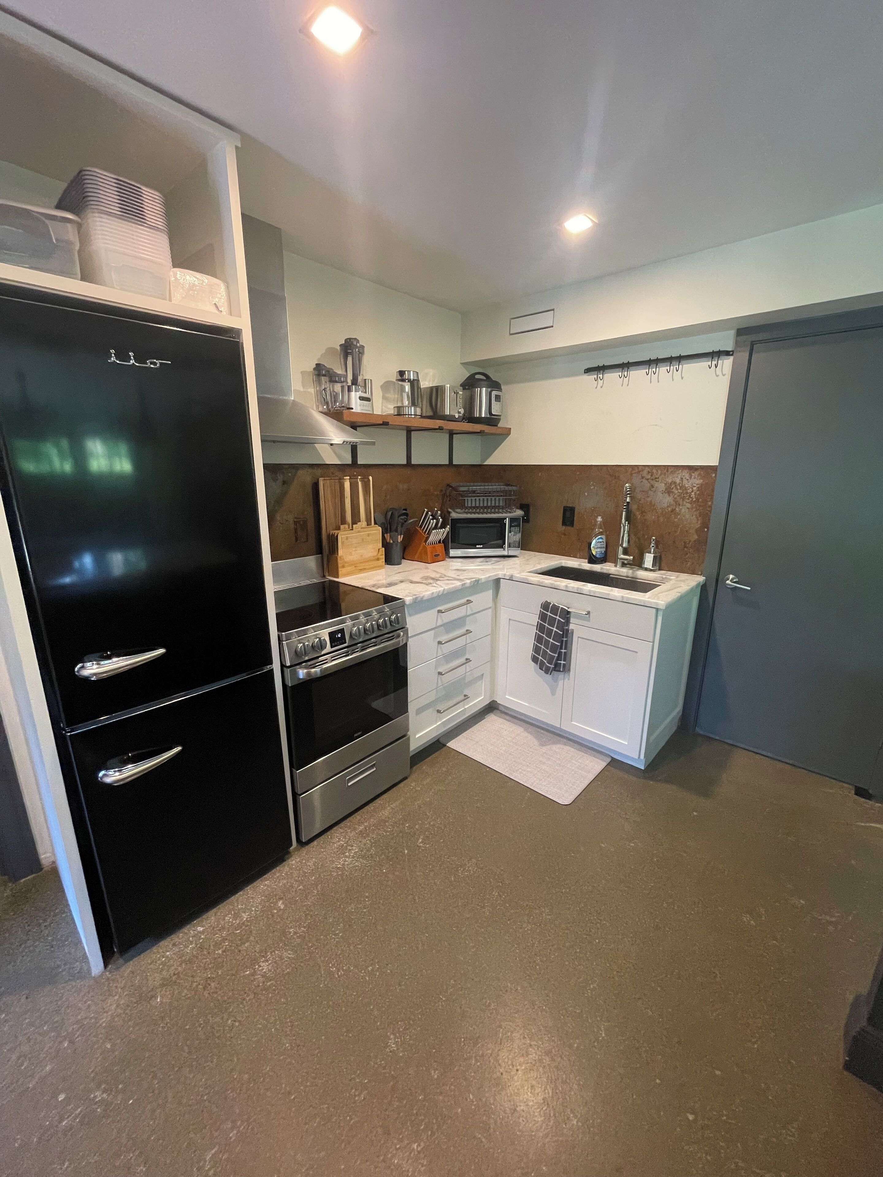 Cocina compacta con refrigerador negro, microondas, estufa y pileta. Hay un estante de madera sobre la estufa y una toalla colgada en la puerta.