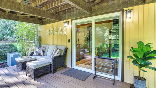 A covered outdoor seating area with wicker furniture and a 'RELAX' sign, a potted plant, and a view of the garden. - Riverfront Sandy Retreat, Fish & Kayak On-Site (Sandy)