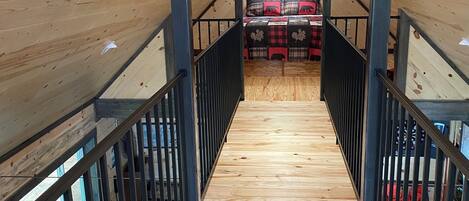 Un interior de madera con una escalera central, una cama con colcha a cuadros rojos y negros, y una alfombra estampada.