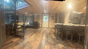 Un intérieur de chalet chaleureux avec un plancher en bois, un coin repas doté de tabourets de bar, une cuisine équipée d’un four à micro-ondes et un escalier en colimaçon menant à une mezzanine.