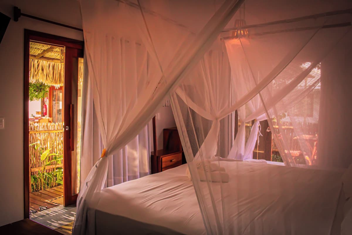Ein Zimmer mit einem Bett unter einem Baldachin, einer Holztür, die nach draußen führt, und einem kleinen Tisch mit einer Lampe.