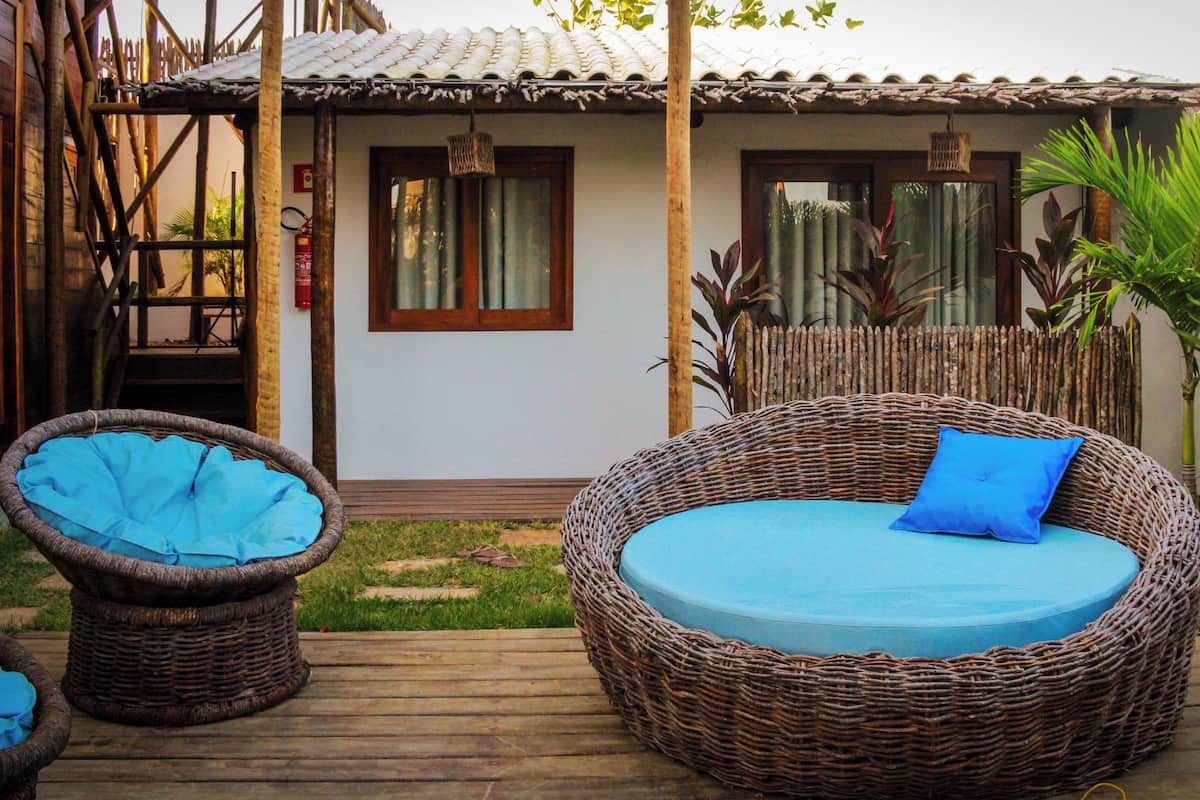 Ein Korbsessel mit blauer Kissenlehne und ein passender Sessel, beide auf einer Holzterrasse vor einem Haus mit Ziegeldach und Bambuszaun.