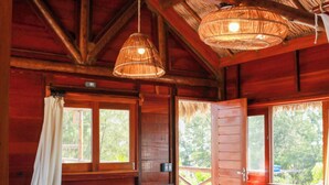A wooden cabin with a thatched roof, a wooden bench with cushions, and a stone floor.