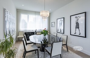 Un comedor moderno con una mesa redonda de cristal, sillas blancas, una araña y cuadros en las paredes.