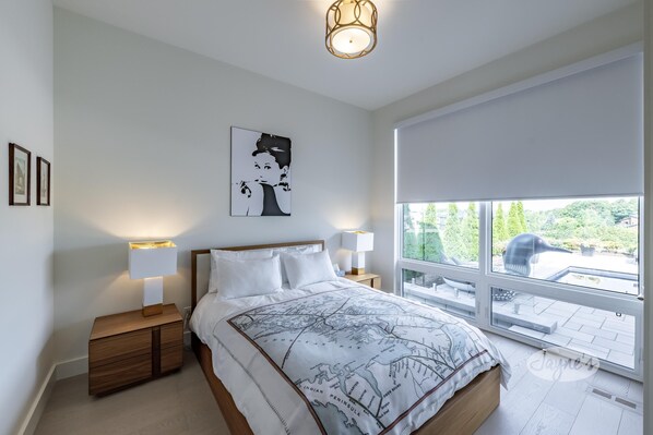 A modern bedroom with a large bed, two bedside tables with lamps, a wall-mounted artwork, and a large window with a view of greenery.