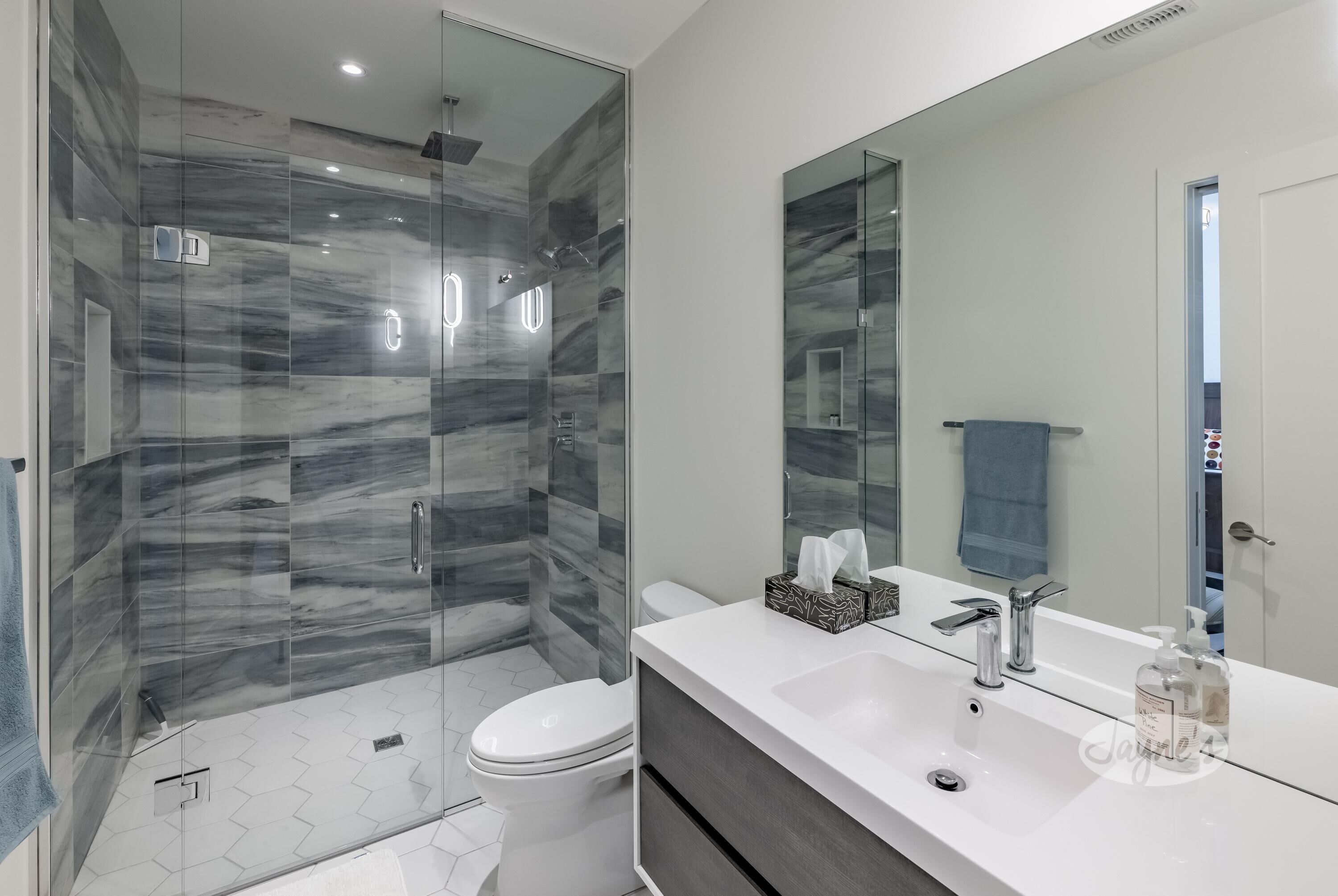 Ein modernes Badezimmer mit einer Glaskabine zur Dusche, einer weißen Toilette und einem großen Spiegel über einem weißen Waschbecken mit Seifenspender.