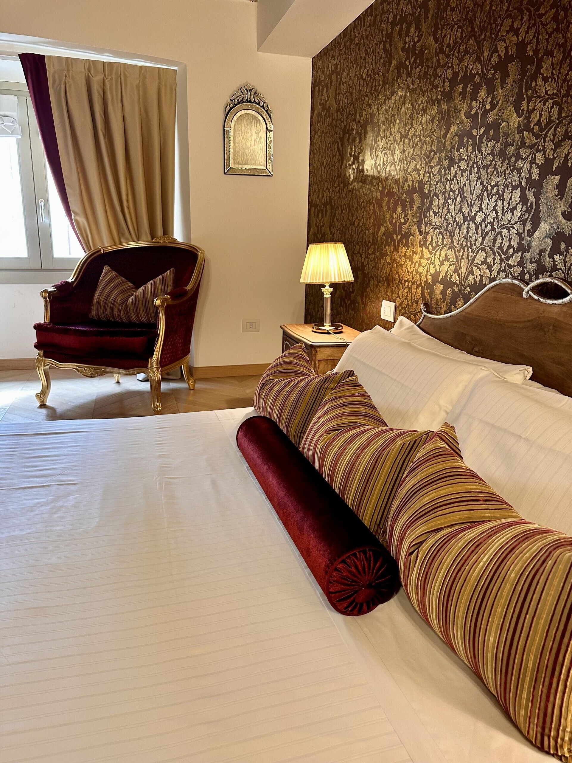 A hotel room with a large bed, a red armchair, a nightstand with a lamp, and a decorative wall panel.