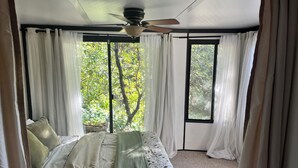 Ein Schlafzimmer mit Bett, weißen Vorhängen, einem Deckenventilator und einem Fenster mit Blick auf Bäume.