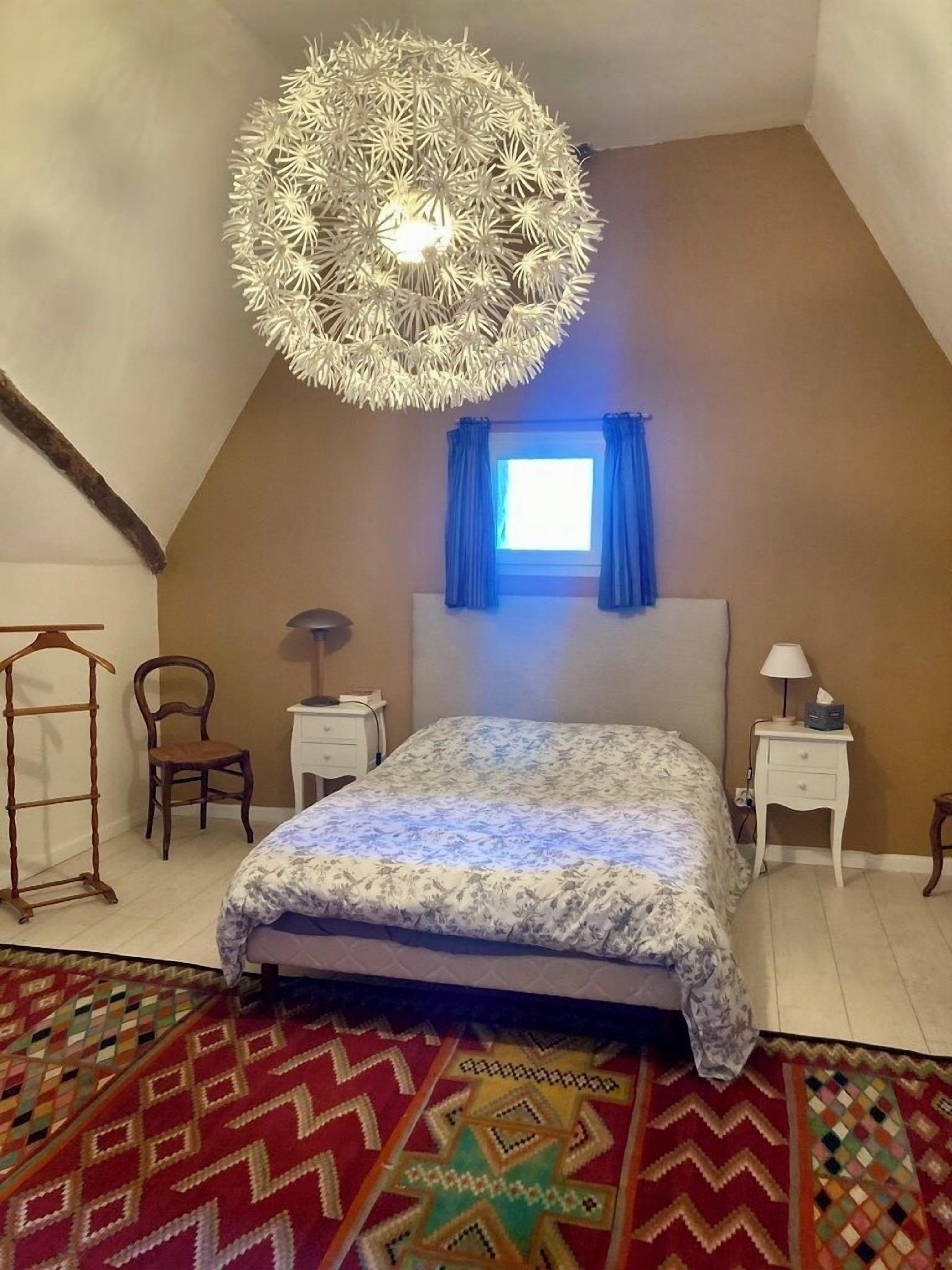 Een slaapkamer met een groot bed, een hanglamp, een raam met gordijnen, een stoel, een nachtkastje en een kamerkleed met een patroon.