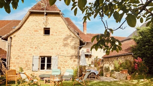 Ein Haus aus Stein mit einem geneigten Dach, einem kleinen Fenster und einer Tür. Ein Garten mit Rasen, einigen Blumentöpfen und Gartenmöbeln.