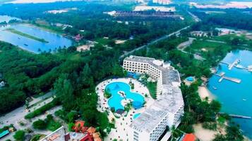 An aerial view of a resort with a large swimming pool, a hotel building, and surrounding greenery.