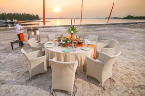 Una mesa redonda sobre la arena de una playa, con sillas de mimbre, un camarero y una vajilla con platos, vasos y un centro de mesa.