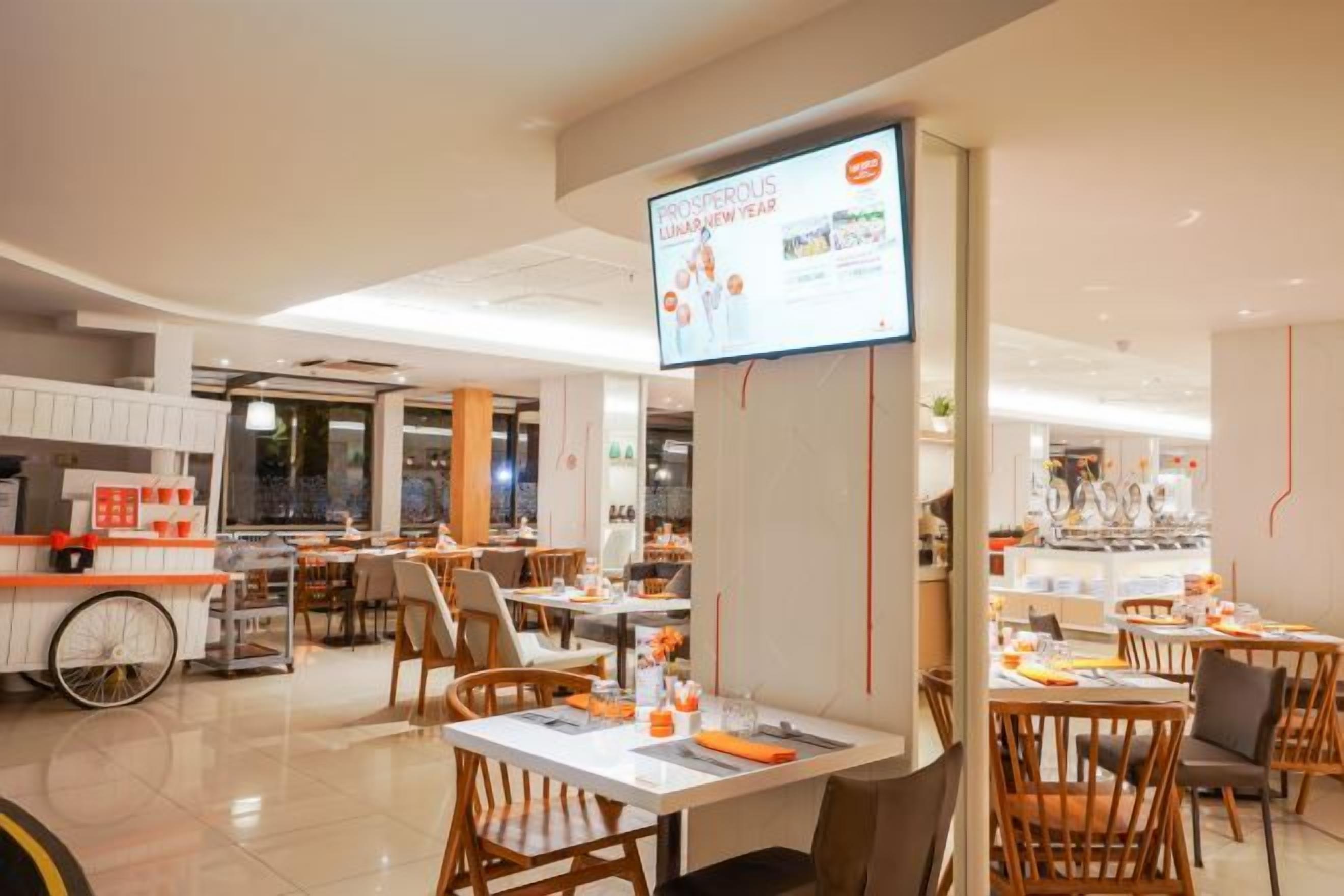 A modern dining area with tables and chairs, a large screen displaying information, and a kitchen area in the background.