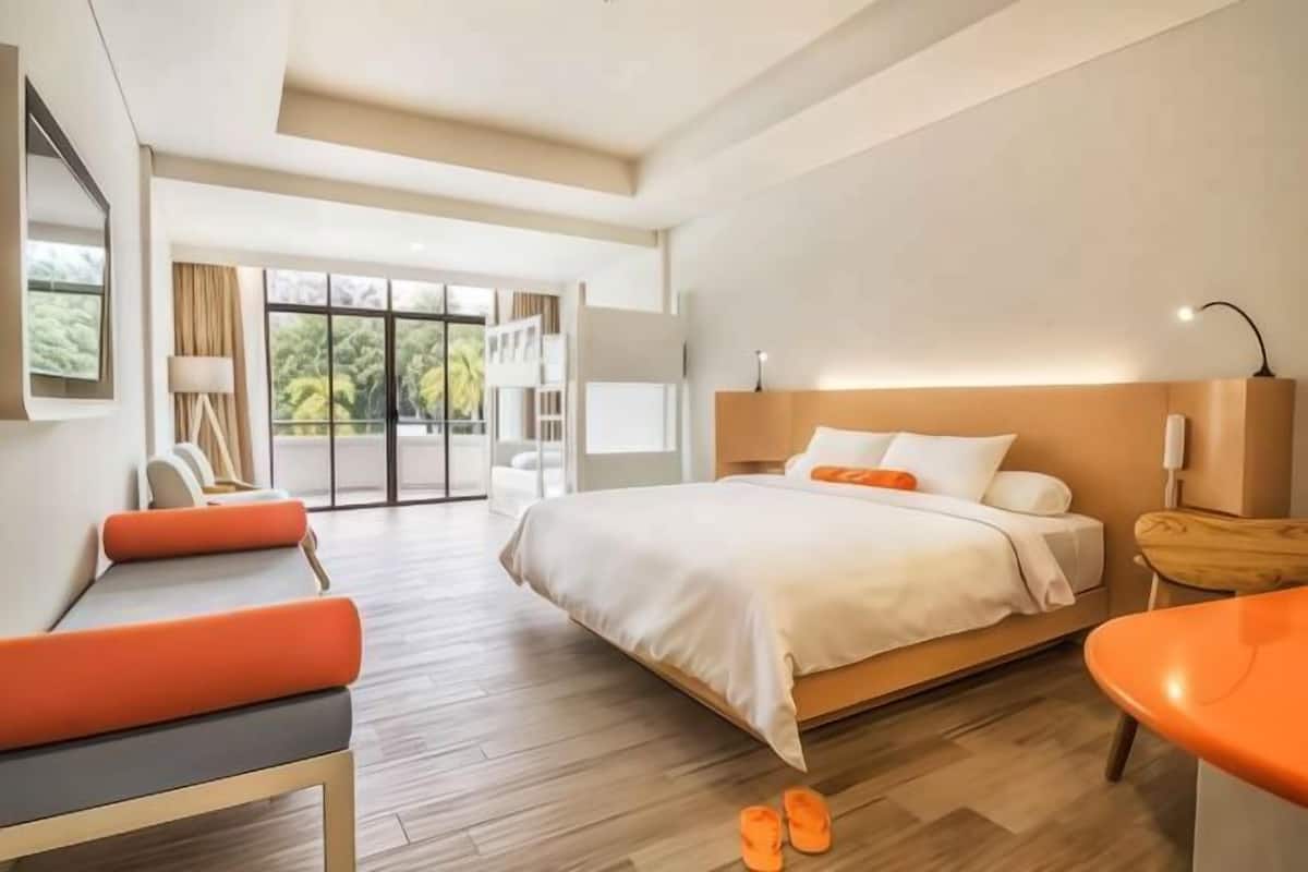 A modern hotel room with a large bed, a seating area with orange cushions, a small table, and a view of greenery outside.
