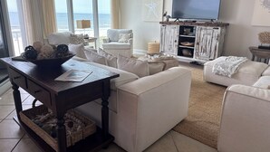 A living room with a large window overlooking the ocean, a television, and a cabinet with decorative items. - BEACHFRONT! OPEN CONCEPT! STUNNING 3 walls of  GLASS! (Pensacola)