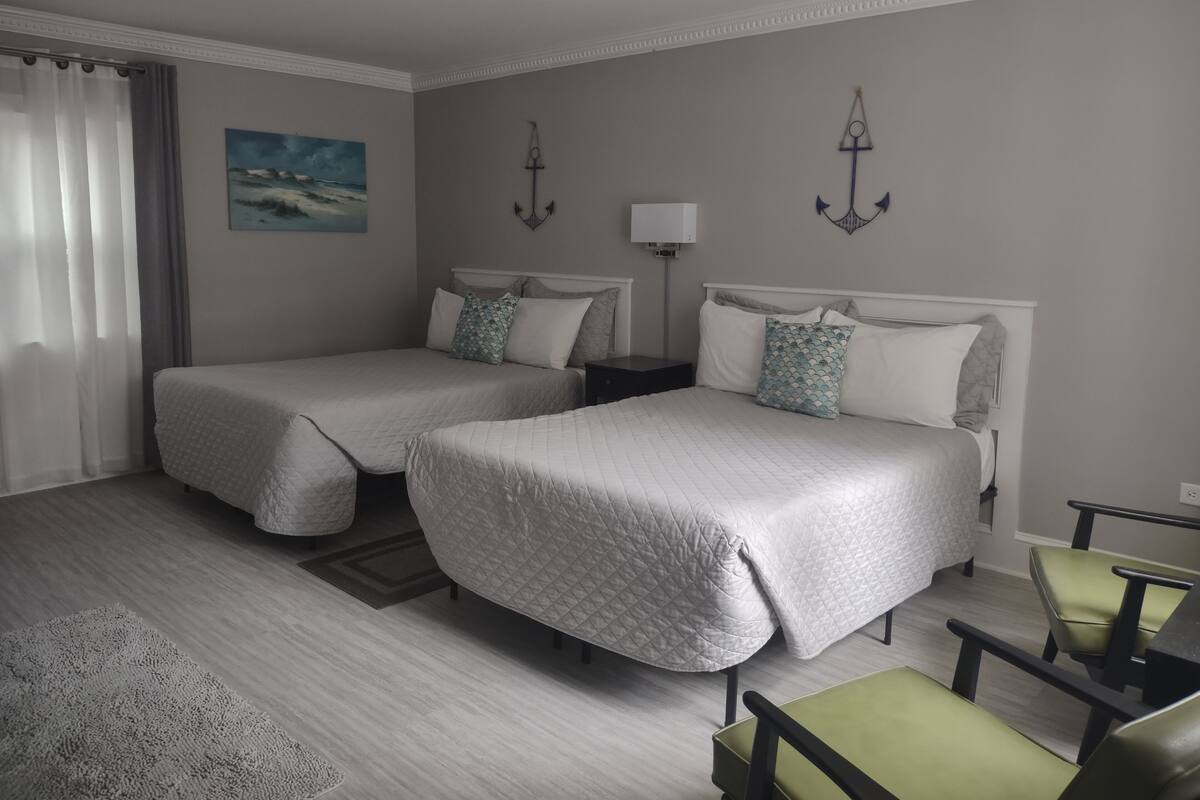 A hotel room with two beds, a nightstand, a lamp, a painting, and a view of the sea.
