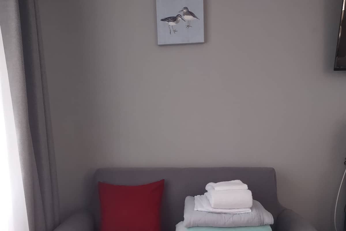 A grey sofa with a red pillow, a stack of folded blankets, and a framed picture on the wall.