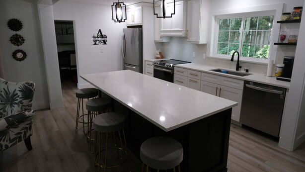 A modern kitchen with a central island, bar stools, a refrigerator, an oven, a dishwasher, and a window with a view of greenery.