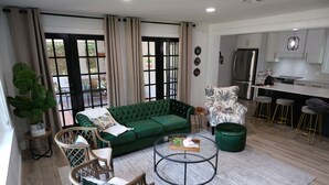 A modern living room with a green sofa, wicker chairs, a round coffee table, and a kitchen area with white cabinets and a stainless steel refrigerator.
