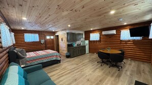 Een blokhut van binnen, met een bed, bank, eettafel, keuken en een televisie aan de wand.