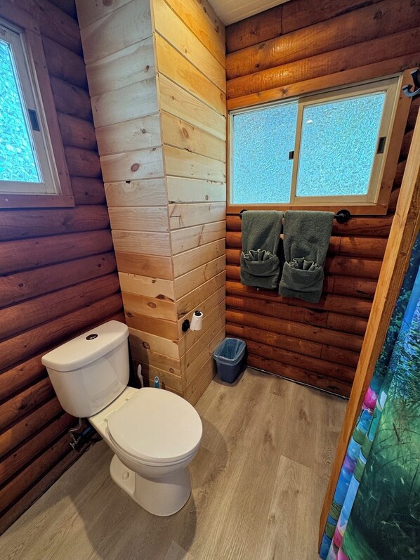 Een houten badkamer in een vakantiehuisje, met een toilet, twee handdoeken aan een rek, en een raam met gezandstraald glas.