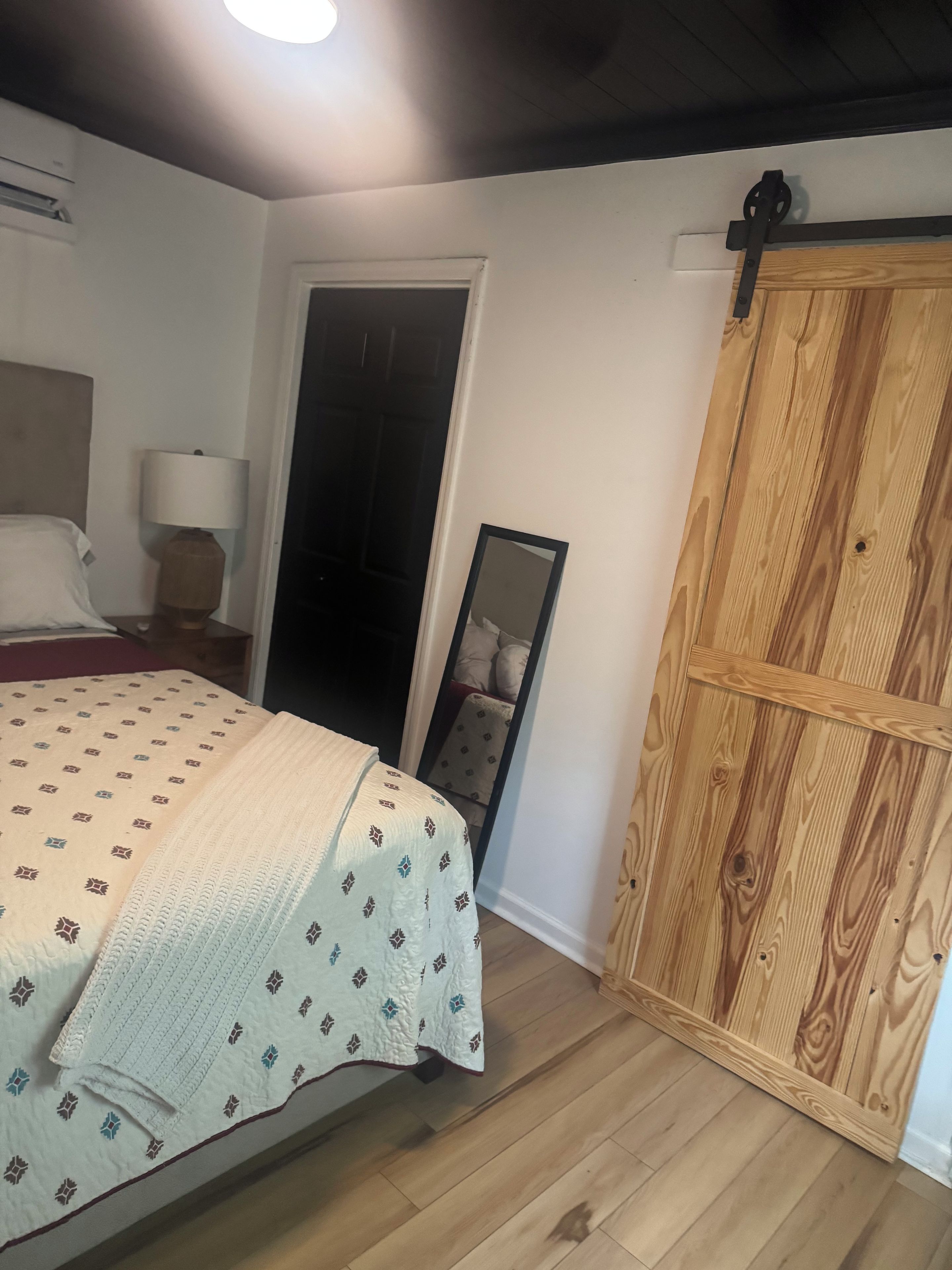 A bedroom with a bed, a nightstand, a lamp, a mirror, and a wooden wardrobe.