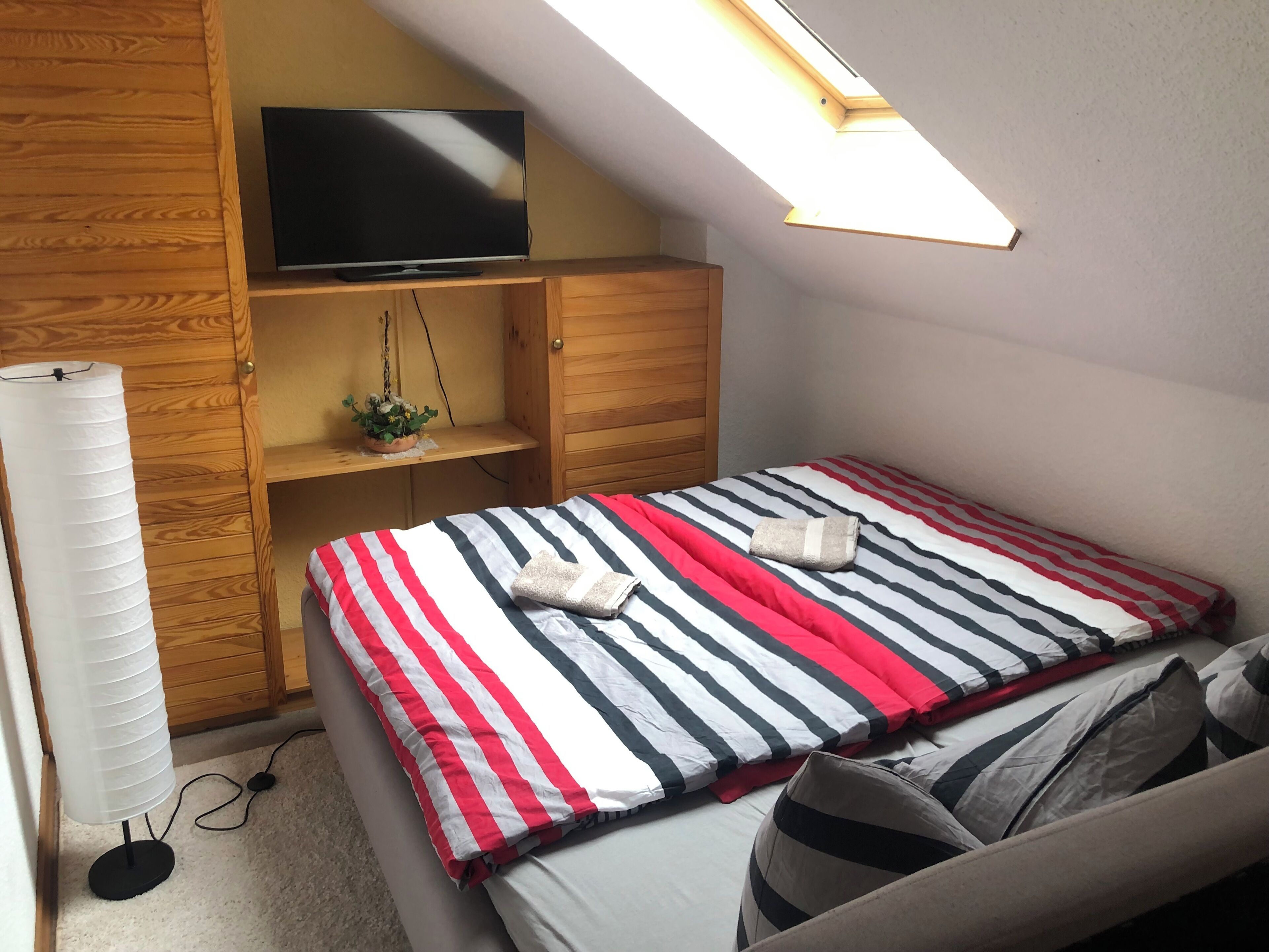 A bedroom with a bed, a TV on a wooden shelf, a skylight, and a fan.