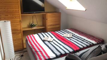 A bedroom with a bed, a TV on a wooden shelf, a skylight, and a fan.