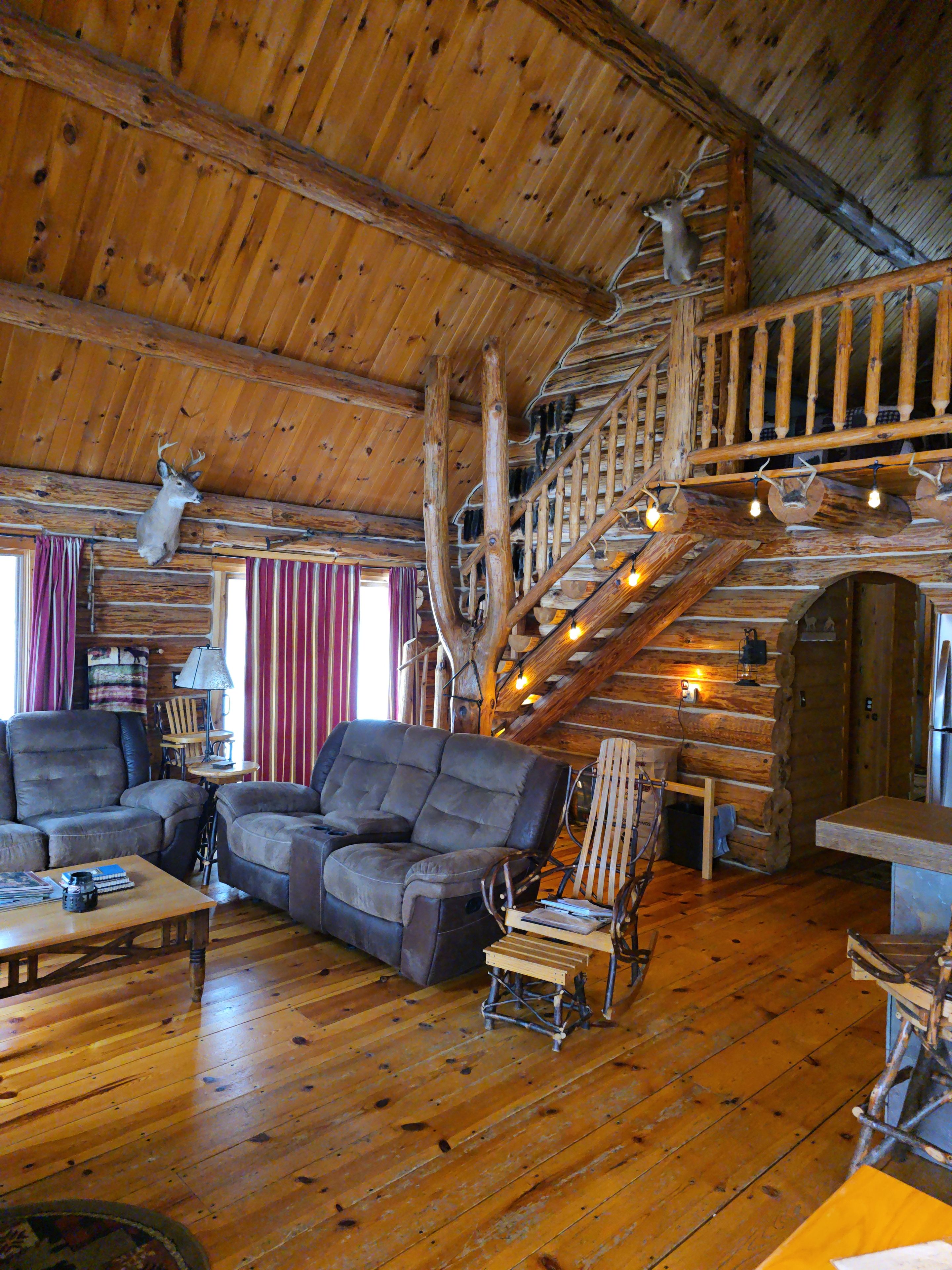 A log cabin interior with a wooden staircase, a fireplace, and a deer head mounted on the wall.