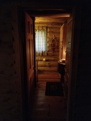 Een smalle houten gang die leidt naar een kamer met een geruit gordijn en een hangende ringlamp.