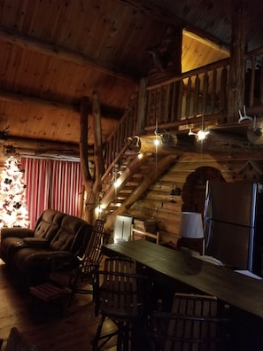 A rustic cabin interior with a wooden staircase, a dining table with chairs, and a leather sofa.