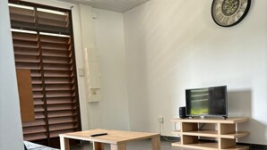 A compact living space with a bed, a small table, a TV on a wooden stand, and a wall clock. - Trésor Caché d'Amazonie Oasis de Calme (Cayenne)